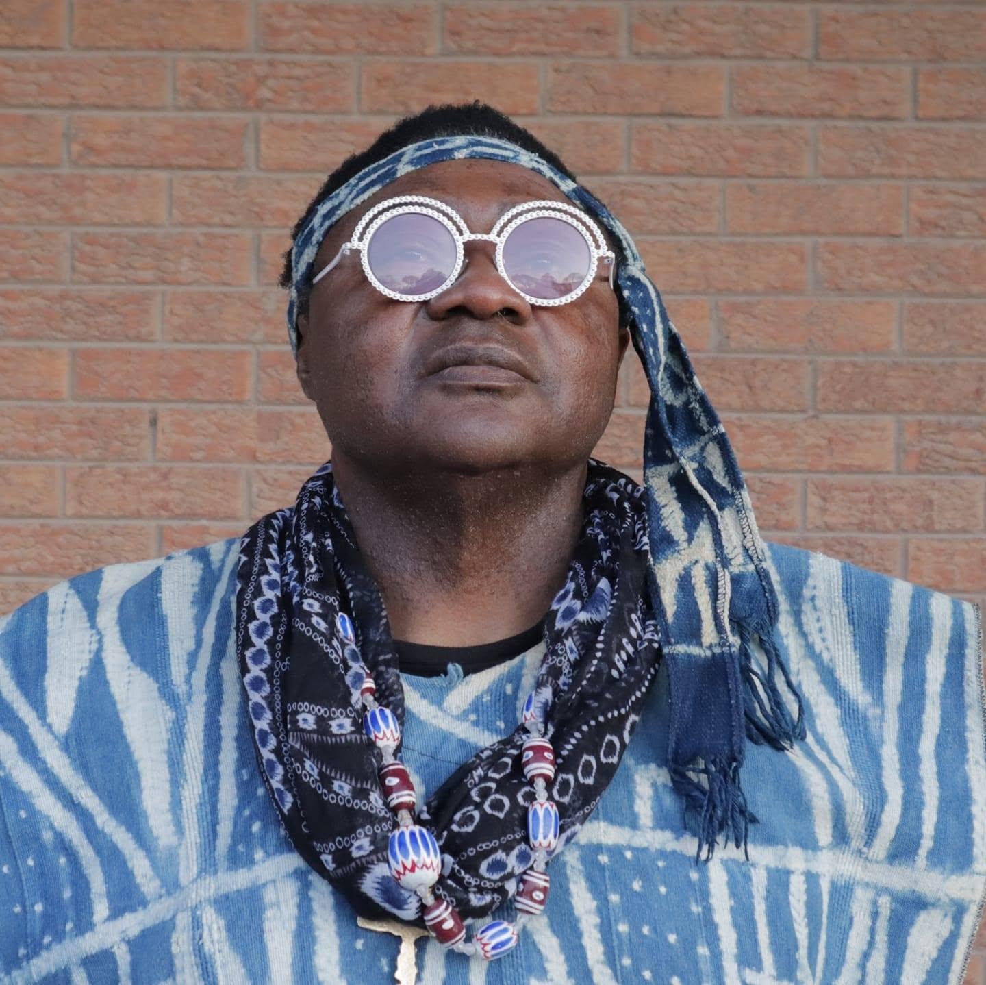 JAH YAMS avec lunettes rondes et bandana motif indigo, devant un mur de briques