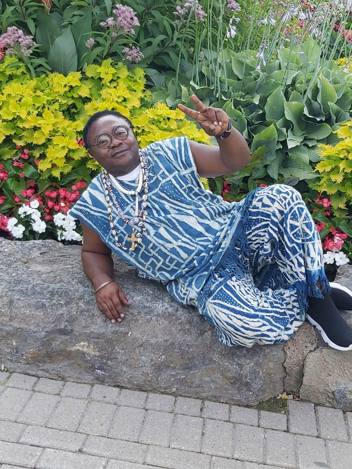 JAH YAMS assis parmi des fleurs, en tenue traditionnelle bleu et blanc, faisant un signe de paix